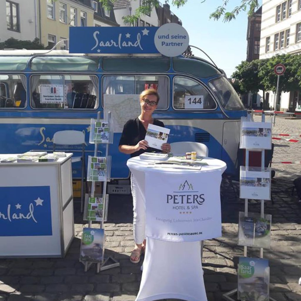 Eine Frau steht an einem Werbestand für Peters Hotel & Spa, der im Freien vor einem blau-weißen Lieferwagen mit der Aufschrift "Saarland" aufgebaut ist. Um sie herum sind auf dem Kopfsteinpflaster Broschüren und Stände aufgebaut.