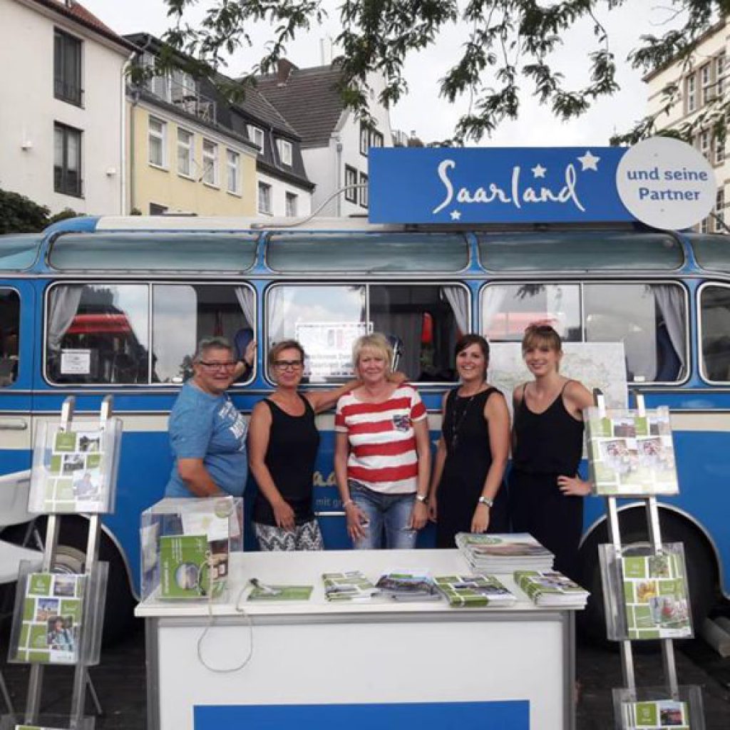 Fünf Personen stehen lächelnd vor einem blau-weißen Oldtimer-Van mit der Aufschrift "Saarland und seine Partner", auf dessen Tisch Broschüren und Flyer für eine Veranstaltung im Freien ausgelegt sind. Im Hintergrund sind Gebäude und Bäume zu sehen.