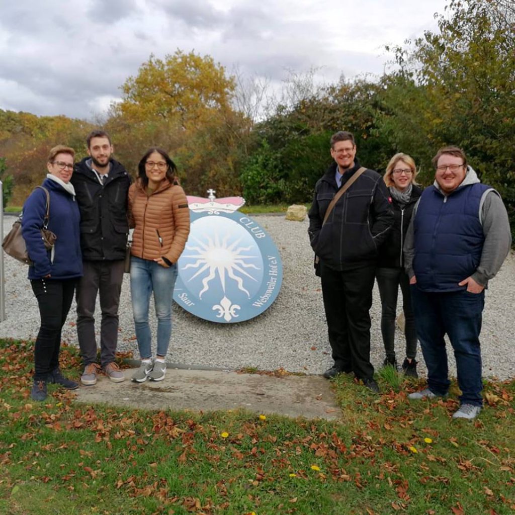 Sechs Personen stehen lächelnd im Freien auf Gras und Kies in der Nähe eines Clubschilds mit einem Sonnen- und Wellenlogo, umgeben von Herbstbäumen unter einem bewölkten Himmel. Sie tragen legere Herbstkleidung und Jacken.