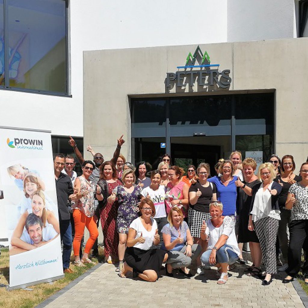 Eine große Gruppe lächelnder Menschen, meist Frauen, posiert gemeinsam vor einem Gebäude mit einem Peters-Schild. Auf der linken Seite steht ein Banner von proWIN International. Die Gruppe ist leger gekleidet und einige zeigen die Daumen nach oben.