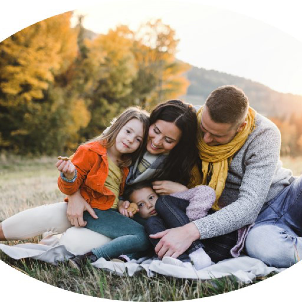 PETERS Kinderkarte 8 Eine vierköpfige Familie sitzt bei Sonnenuntergang auf einer Decke in einem grasbewachsenen Feld. Sie lächeln und umarmen sich gegenseitig, umgeben von herbstlichen Bäumen und Hügeln im Hintergrund.