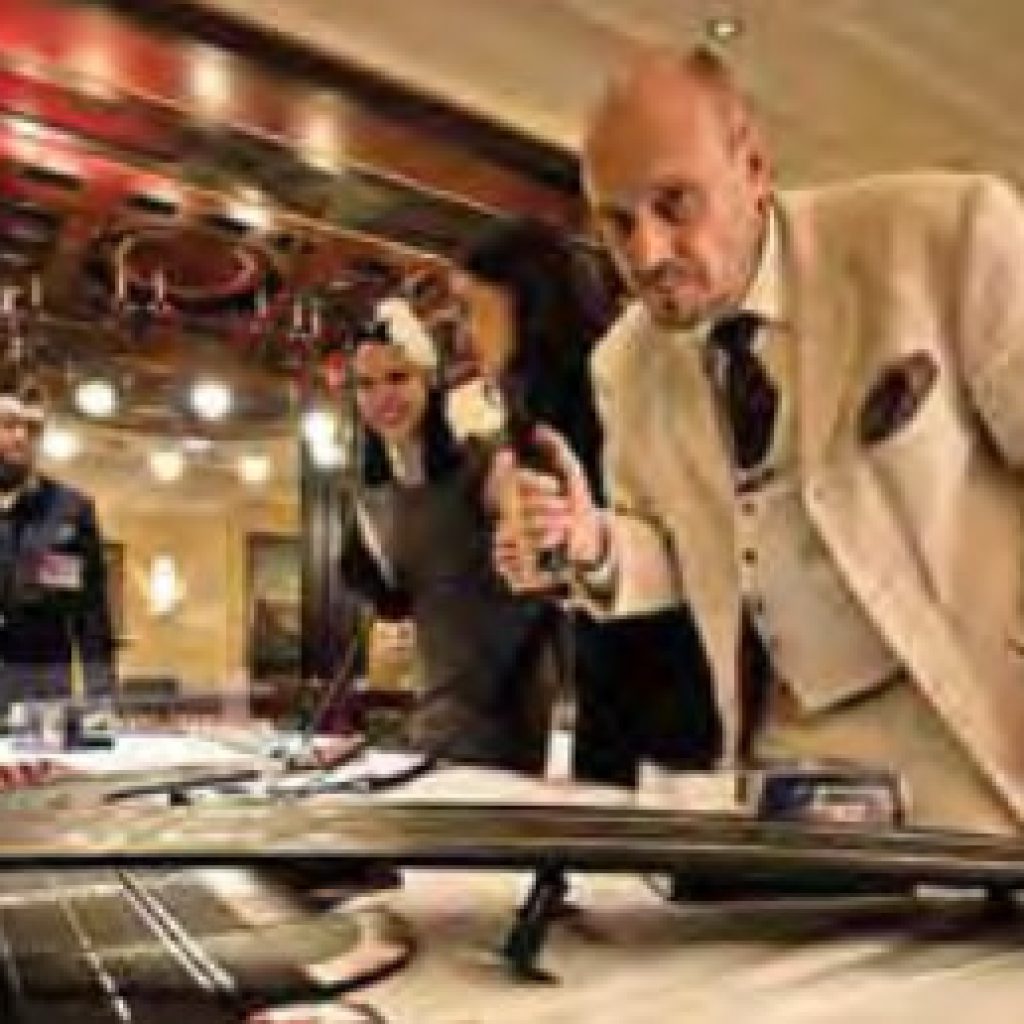 Ein Mann in einem cremefarbenen Anzug beugt sich über einen Tisch, hält ein Mikrofon in der Hand und untersucht eine Slotcar-Rennbahn, während zwei Personen zusehen und ein weiterer Mann in der Nähe in einem schwach beleuchteten Raum steht.