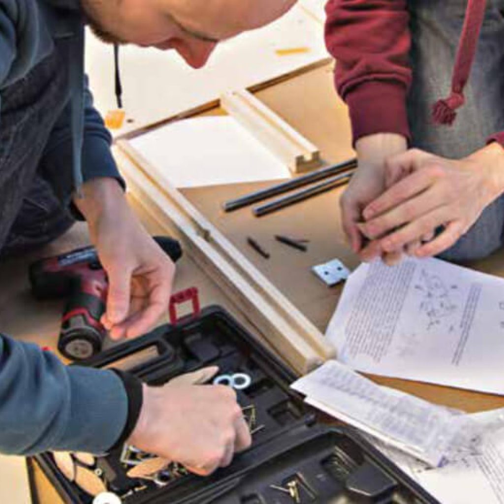 Zwei Personen, die mit Hilfe von Werkzeugen und Anleitungen Möbel zusammenbauen; die eine greift in einen Werkzeugkasten, während die andere die Beschläge hält, während die Montageteile und Unterlagen auf einem Tisch ausgebreitet sind.