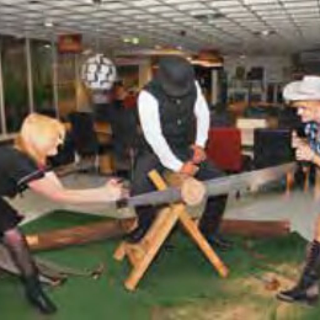 Drei kostümierte Personen benutzen eine große Säge für zwei Personen, um einen Baumstamm im Haus zu sägen. Eine Person steht in der Mitte und schaut zu, während die beiden anderen, die mit Stiefeln und Hüten bekleidet sind, die Säge an den gegenüberliegenden Enden bedienen.