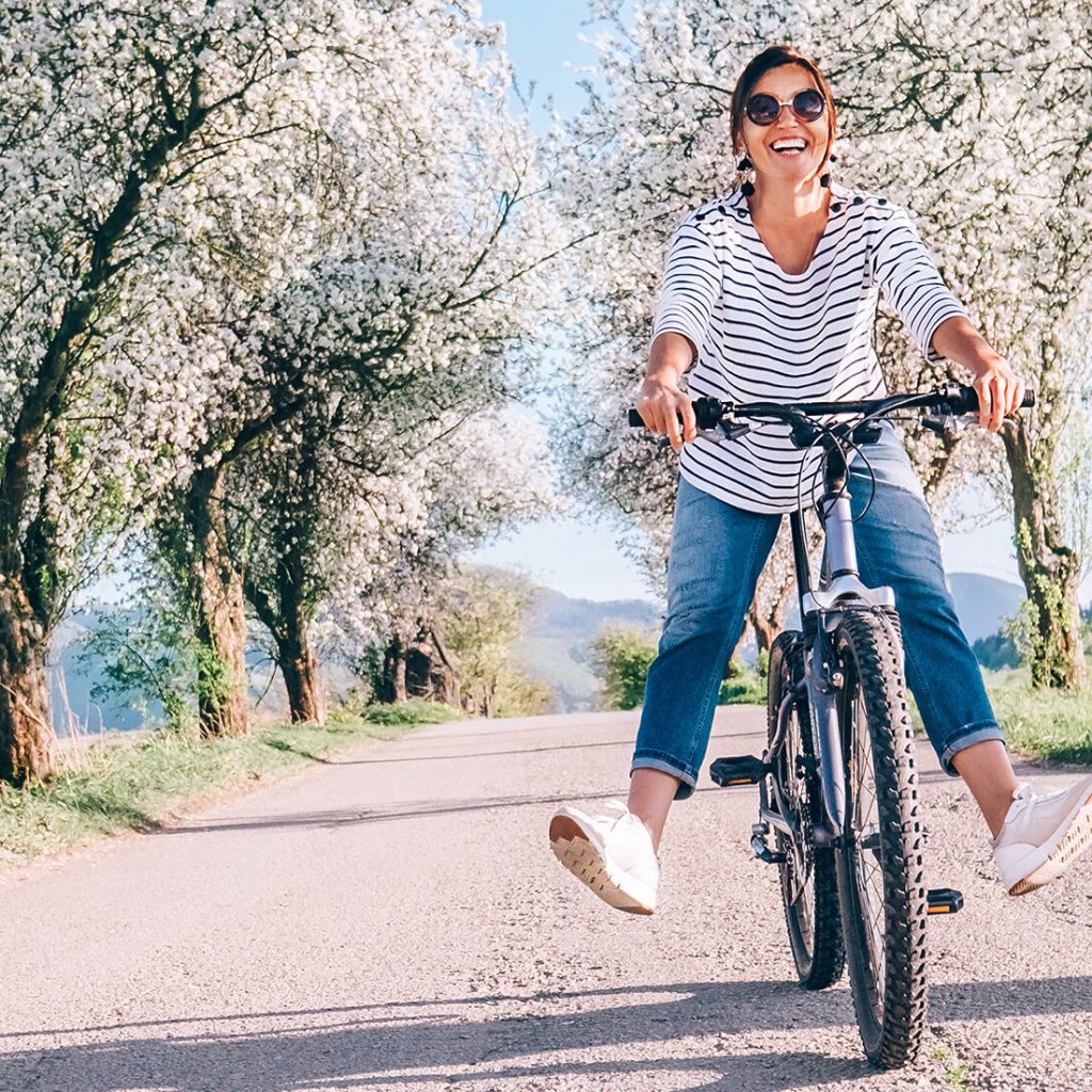 Der Frühling kehrt ein 11 Eine Frau fährt lächelnd und mit ausgestreckten Beinen auf einer sonnigen Straße, die von blühenden Bäumen gesäumt ist, mit dem Fahrrad.