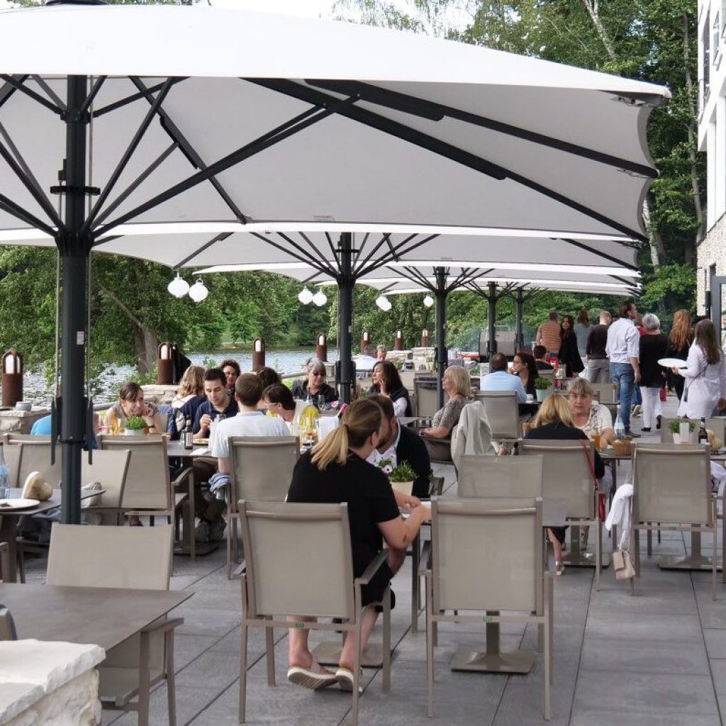 Sommer-Events bei Peter's Hotel & Spa 5 Menschen speisen im Freien unter großen weißen Sonnenschirmen auf einer Terrasse am Flussufer. Die Szene ist lebhaft, die Gäste sitzen an Tischen, genießen Speisen und Getränke, umgeben von Grün und Wasser.