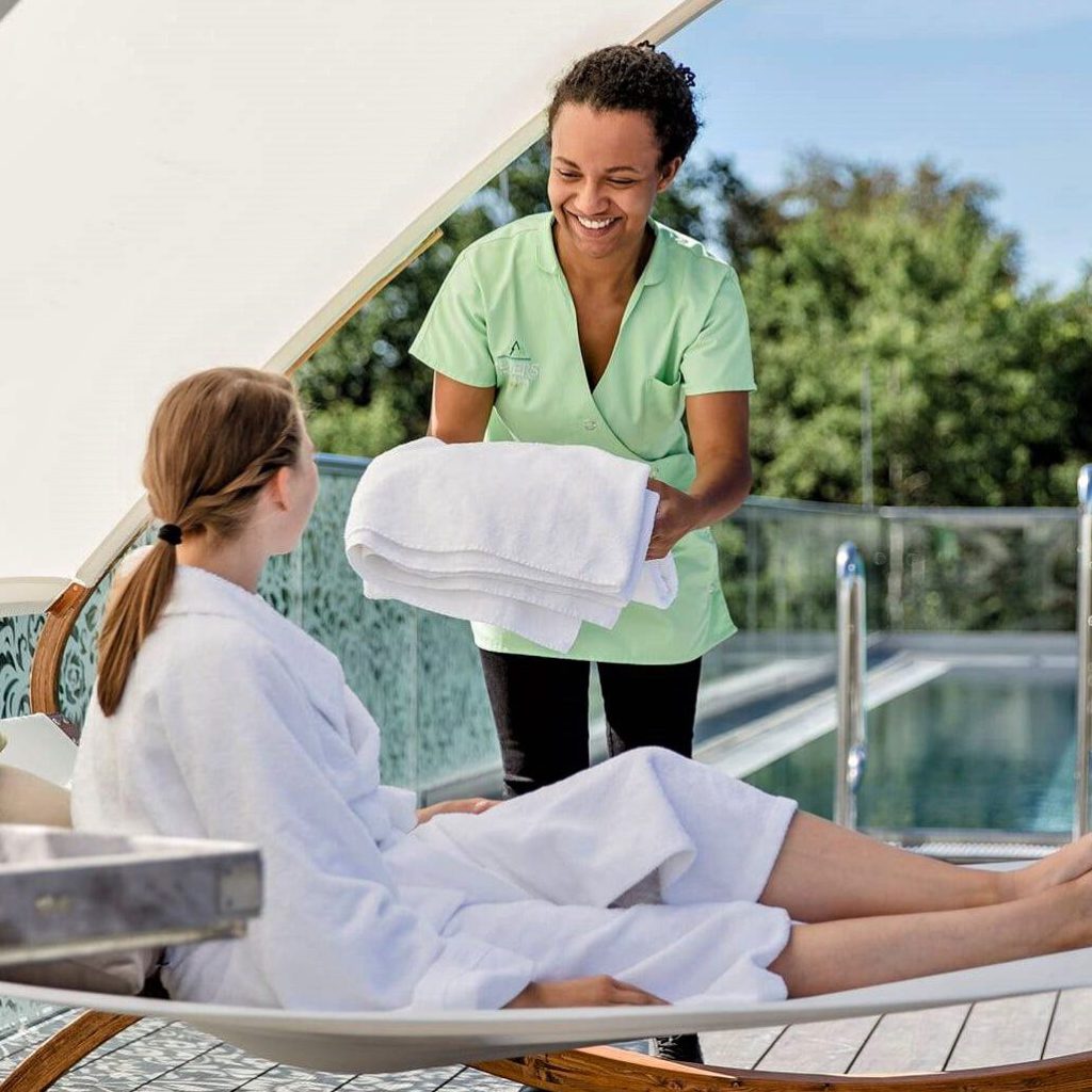 Gastfreundschaft wird bei uns groß geschrieben 2 Ein lächelnder Mitarbeiter reicht einer Frau, die sich auf einem Liegestuhl an einem Außenpool entspannt, frische Handtücher. Im Hintergrund sind Bäume und ein Glasgeländer zu sehen.