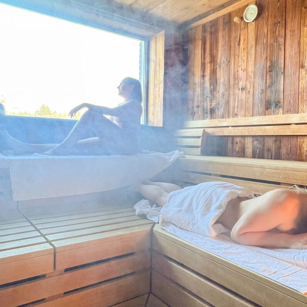 Es ist herbstlich - Jetzt Immunsystem stärken! 10 Drei Personen entspannen sich in einer hölzernen Sauna, die mit Dampf gefüllt ist; zwei sitzen auf der oberen Bank in der Nähe eines großen Fensters, während eine Frau auf einer unteren Bank liegt, alle in Handtücher gehüllt. Das Sonnenlicht fällt von draußen herein.