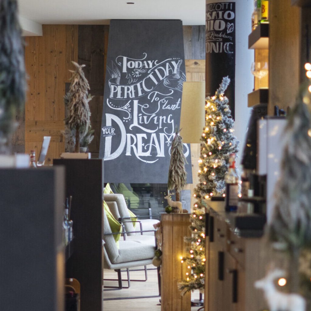 Der Winter ist da 9 Ein gemütliches, warm beleuchtetes Café, dekoriert mit kleinen, beleuchteten Weihnachtsbäumen und rustikalen Holzakzenten. Auf einer Kreidetafel steht: "Heute ist der perfekte Tag, um deinen Traum zu leben".