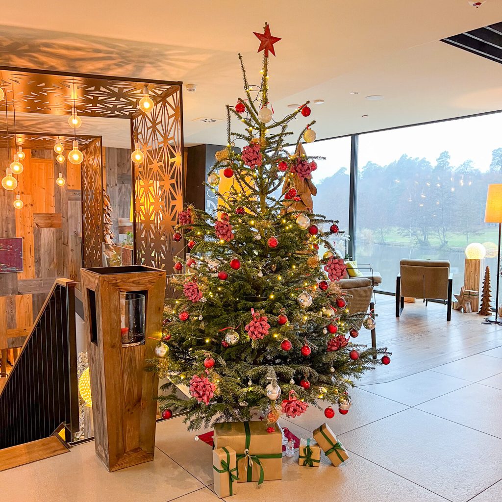 Rituale an Weihnachten 8 Ein geschmückter Weihnachtsbaum mit rotem Schmuck, Kugeln, Tannenzapfen und einer roten Sternspitze steht drinnen auf einem Kachelboden, umgeben von eingepackten Geschenken. Im Hintergrund sind eine warme Beleuchtung und große Fenster mit Blick nach draußen zu sehen.