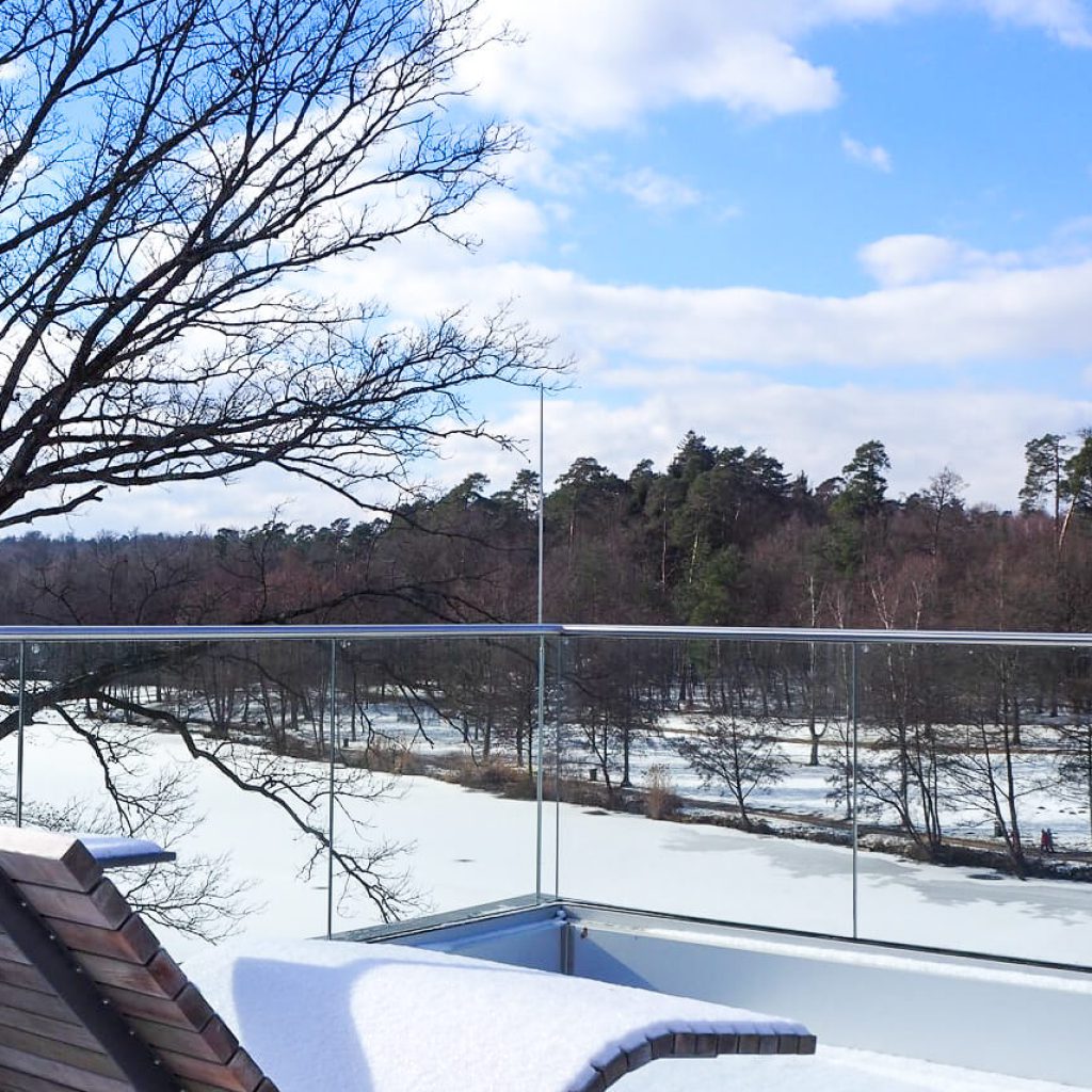Die Naherholung in Jägersburg 7 Ein verschneiter Balkon mit einem hölzernen Liegestuhl bietet einen Blick auf einen zugefrorenen Fluss und eine Landschaft mit blattlosen Bäumen unter einem teilweise bewölkten blauen Himmel.