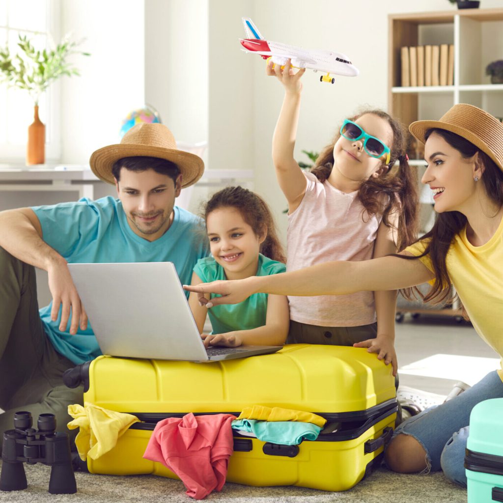 Eine fröhliche Familie mit zwei Kindern sitzt in einem hellen Wohnzimmer, umgeben von gepackten Koffern. Die Eltern schauen auf einen Laptop, während ein Kind ein Spielzeugflugzeug hält und das andere lächelt. Kleidung und Reiseutensilien liegen verstreut herum.