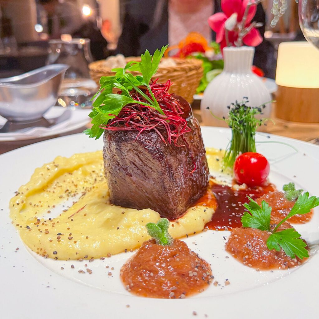 Eine Genussvolle Reise durch den Saar-Pfalz-Kreis 5 Ein Gourmet-Gericht mit einem gebratenen Rinderfilet auf cremigem Kartoffelpüree, garniert mit Kräutern, Kirschtomaten und kunstvoll platzierten Saucen auf einem weißen Teller, angerichtet auf einem eleganten Restauranttisch.