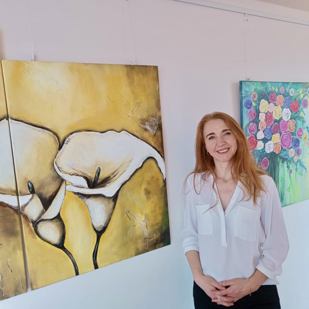 Kunstwerke von Malgorzata Weingardt-Lipowczan 4 Eine Frau mit langen roten Haaren, weißer Bluse und schwarzer Hose, steht lächelnd neben zwei farbenfrohen Gemälden an einer Wand - eines mit Calla-Lilien auf gelbem Hintergrund und eines mit einem Blumenstrauß auf grünem Hintergrund.