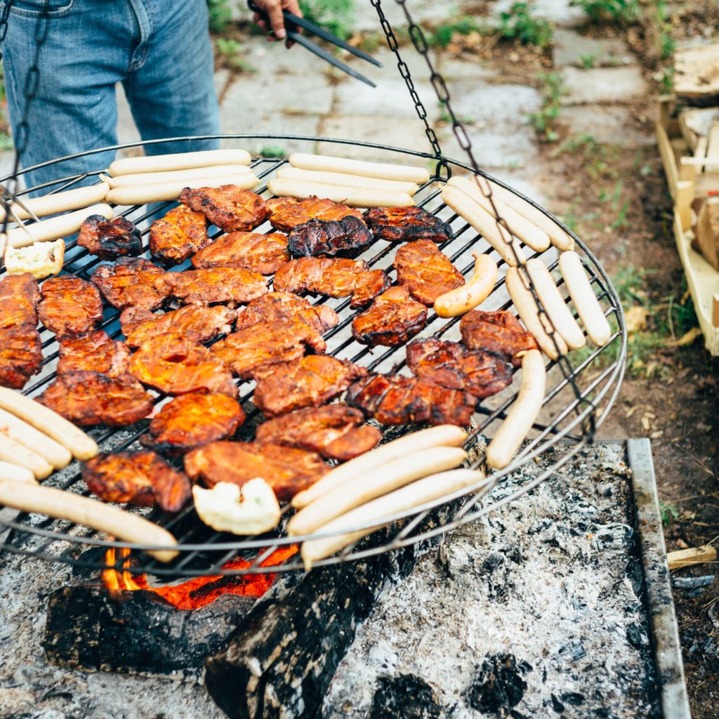 BBQ nach traditionell saarländischer Art 3 Eine Person grillt marinierte Hähnchenteile und Weißwürste auf einem runden, aufgehängten Metallgrill über einem offenen Feuer im Freien, in dessen Nähe gehacktes Holz gestapelt ist.