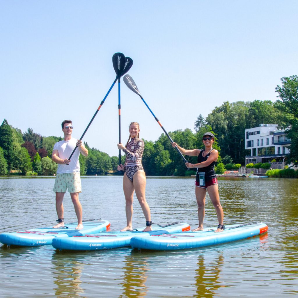 Der perfekte Sommertag am See 2 Drei Personen stehen auf Paddelbrettern in einem ruhigen See und halten ihre Paddel in einer Dreiecksform zusammen. Im Hintergrund sind Bäume und ein weißes Gebäude unter einem klaren blauen Himmel zu sehen.