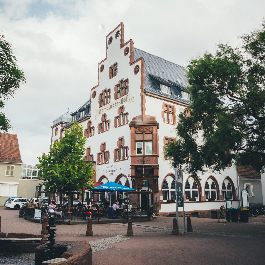 Ein historisches weißes Gebäude mit verschnörkelten Giebeln und roten Verzierungen steht auf einem Platz mit Kopfsteinpflaster. Die Menschen sitzen in einem Café im Freien. Bäume und andere Geschäfte umgeben den Platz an einem teilweise bewölkten Tag.