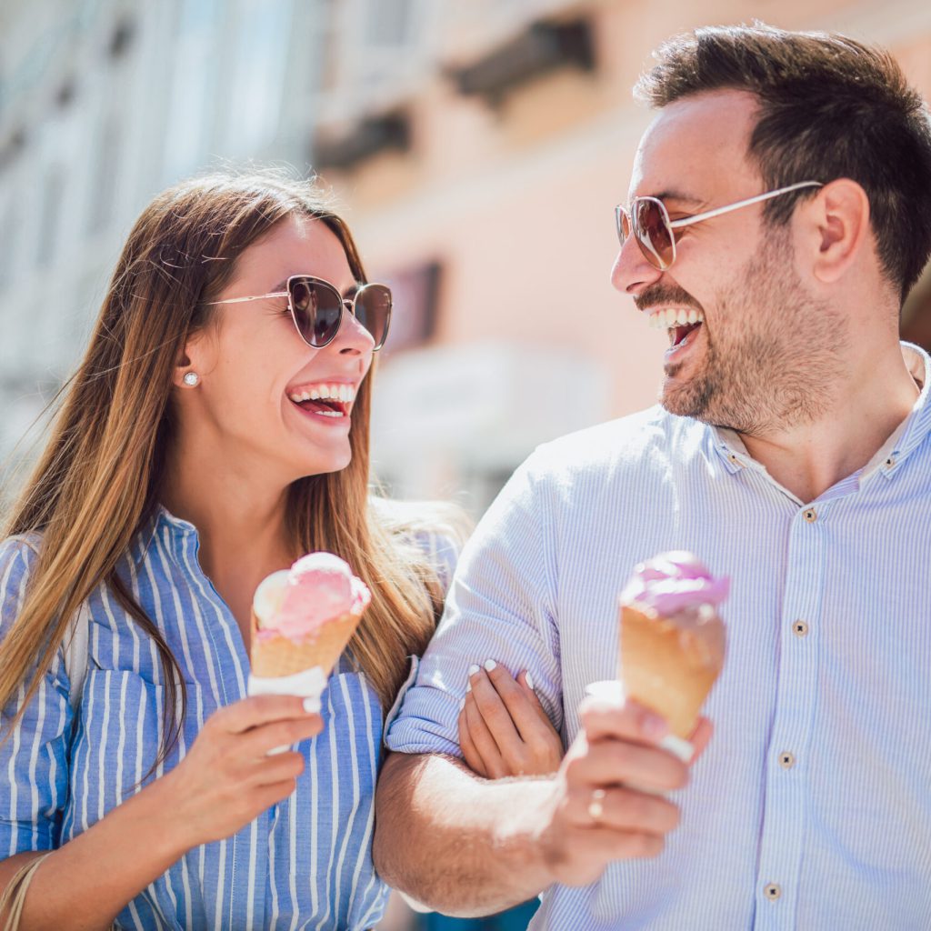 Ein lächelnder Mann und eine Frau mit Sonnenbrille genießen Eiswaffeln, während sie an einem sonnigen Tag im Freien spazieren gehen und sich glücklich ansehen.