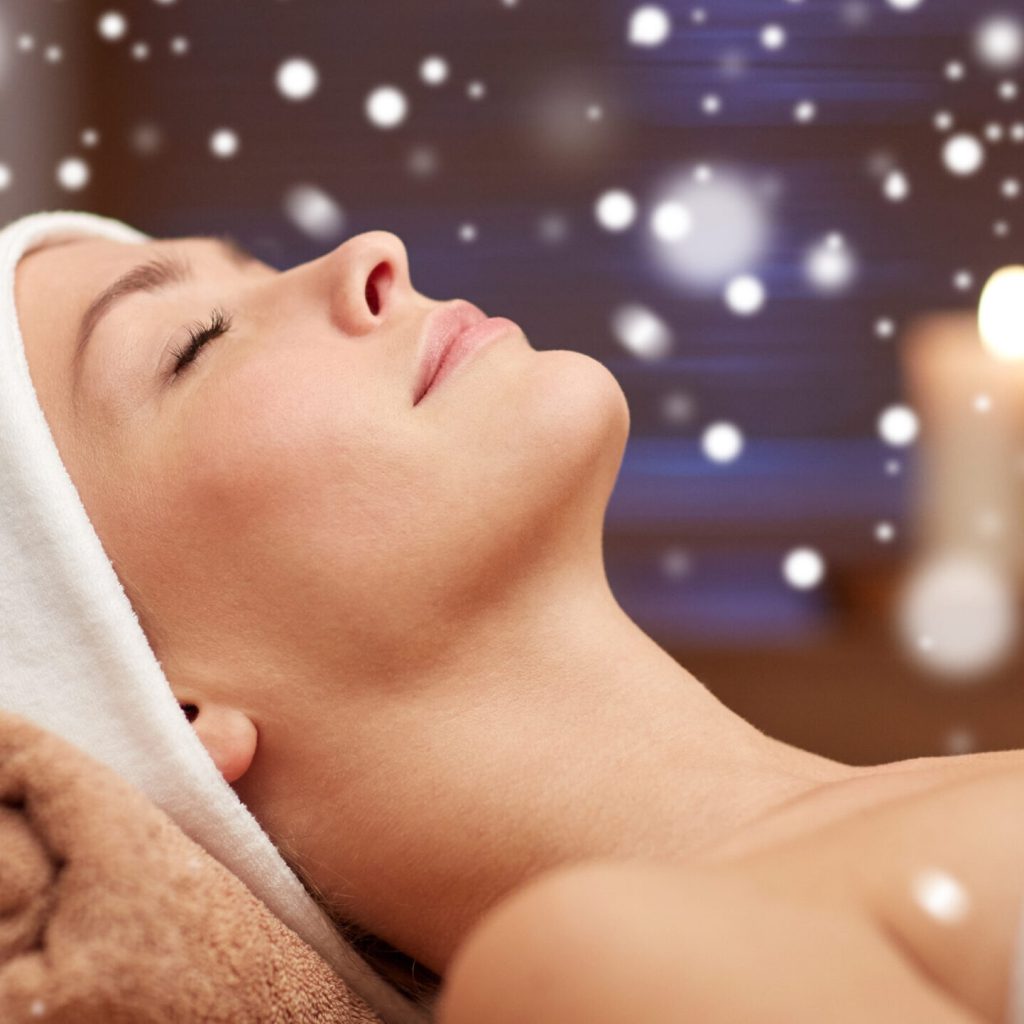 Winterwellness: Stärken Sie Ihr Immunsystem in unserer exklusiven Spa-Oase 1 Eine Frau mit Stirnband entspannt sich mit geschlossenen Augen auf einer Wellnessliege, umgeben von weichen Handtüchern und leuchtenden Kerzen, während weiße Bokeh-Lichter, die an Schneeflocken erinnern, umherschweben.