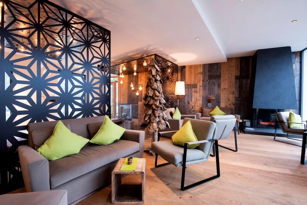 Modernes Wohnzimmer mit braunen Sofas und grünen Kissen, gemustertem schwarzen Raumteiler, Holzboden, kleinem Tisch, dekorativer Beleuchtung und großen Fenstern, die natürliches Licht hereinlassen.