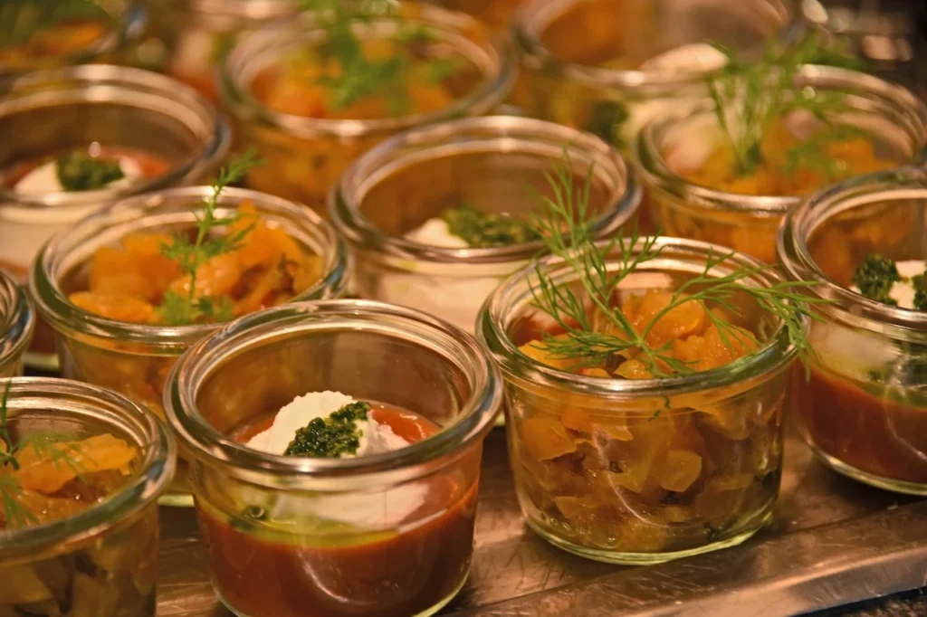 Mehrere kleine Gläser, gefüllt mit bunten Häppchen, belegt mit frischem Dill, Sahne und grüner Pestosauce, dicht an dicht auf einem Tablett angeordnet.