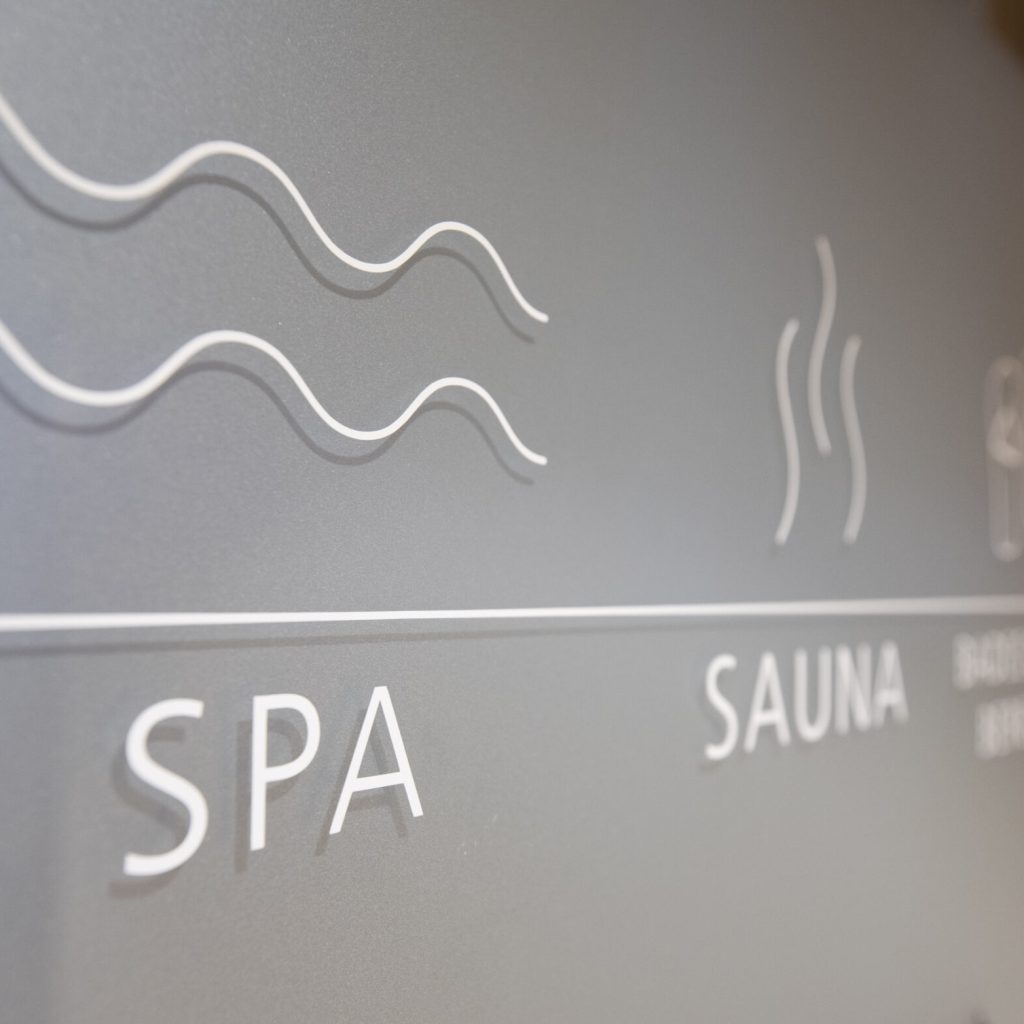 Winterliche Hautpflege: Verwöhnprogramm für Körper und Seele 16 Nahaufnahme eines Milchglasschilds mit minimalistischen Strichsymbolen für ein Spa und eine Sauna, begleitet von den Worten "SPA" und "SAUNA" in weißer Schrift.