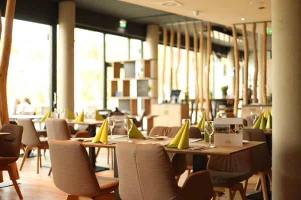 Das moderne Restaurant-Interieur im PETERS Hotel & Spa Saarland besticht durch Holztische und gepolsterte Stühle. Jeder Tisch ist mit grünen, gefalteten Servietten und Gläsern gedeckt. Große Fenster füllen den Raum mit natürlichem Licht, ergänzt durch dekorative Holzelemente an den Wänden.