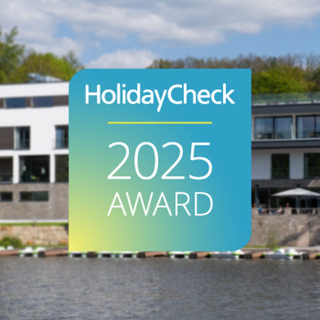 Ein modernes Hotel am See mit Bäumen im Hintergrund, überlagert von einem großen HolidayCheck 2025 Award-Abzeichen in blauen, grünen und gelben Farbtönen in der Mitte des Bildes.