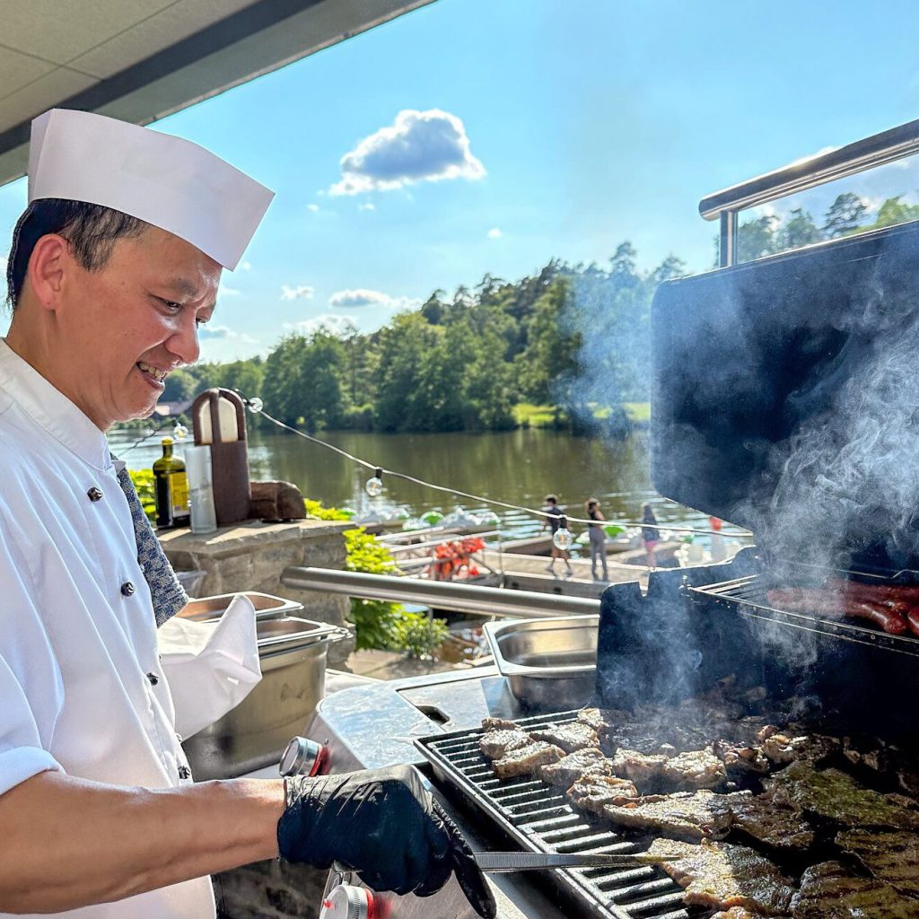 Kulinarische Hochgenüsse unter freiem Himmel 13 Ein Koch in weißer Uniform und Mütze grillt lächelnd Fleisch auf einem Grill im Freien, mit malerischem Blick auf den See, Bäume und blauen Himmel im Hintergrund. Dampf steigt vom Grill auf.