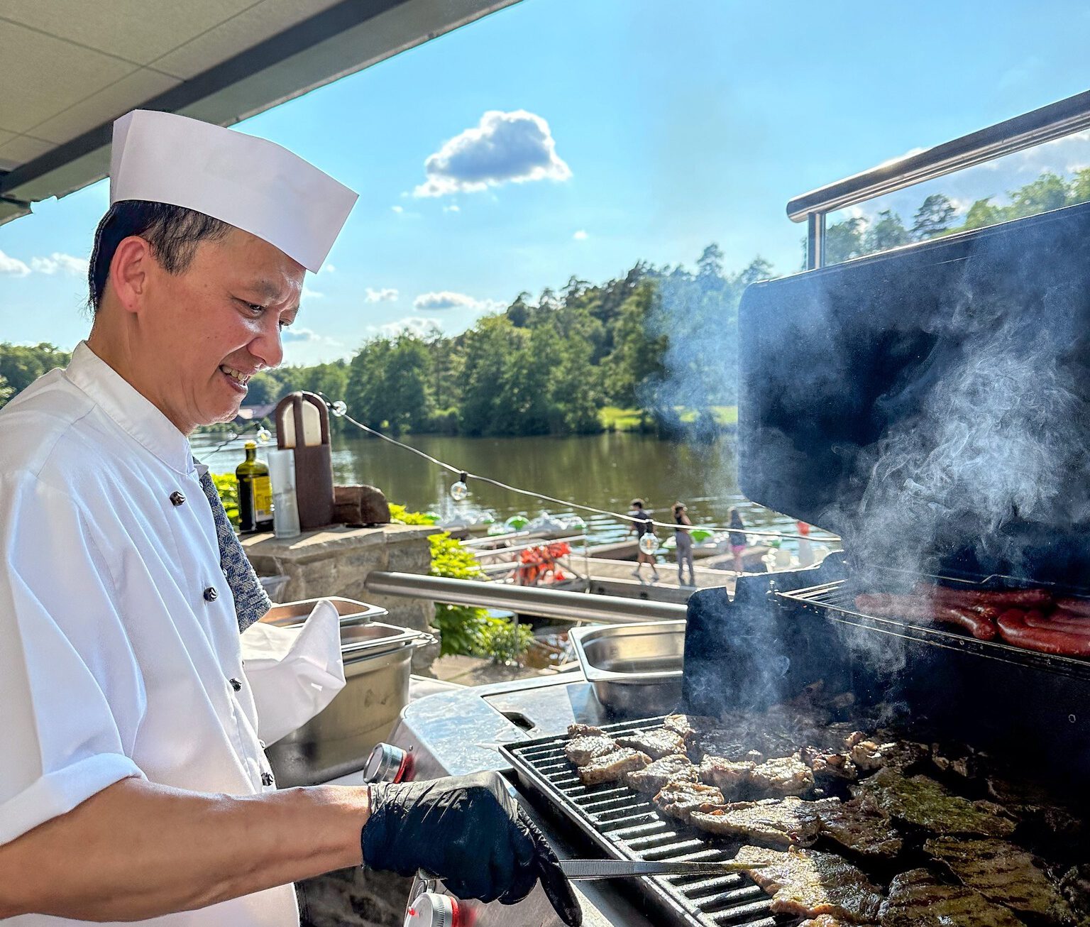 Stories - unser Hotel- & Wellness-Blog 12 Ein Koch in weißer Uniform und Mütze grillt lächelnd Fleisch auf einem Grill im Freien, mit malerischem Blick auf den See, Bäume und blauen Himmel im Hintergrund. Dampf steigt vom Grill auf.