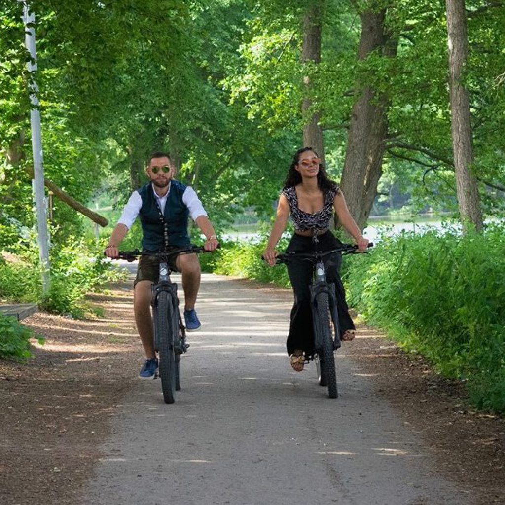 Zwei Personen fahren an einem sonnigen Tag nebeneinander mit dem Fahrrad auf einem von Bäumen gesäumten Weg in einem Park, umgeben von üppigem Grün.