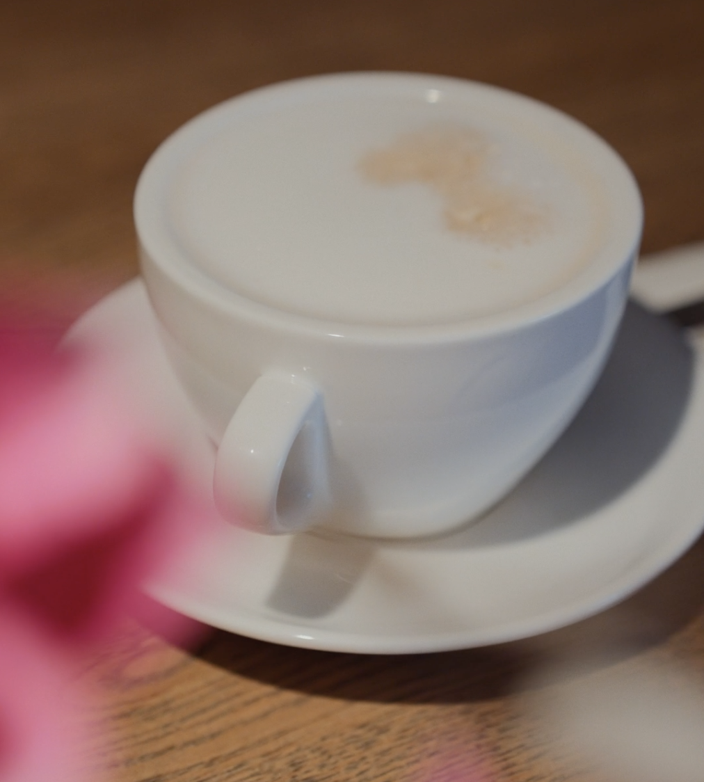 Eine weiße Tasse, gefüllt mit einem schaumigen Kaffeegetränk, steht auf einer passenden Untertasse auf einem Holztisch, mit einem verschwommenen rosa Objekt im Vordergrund.
