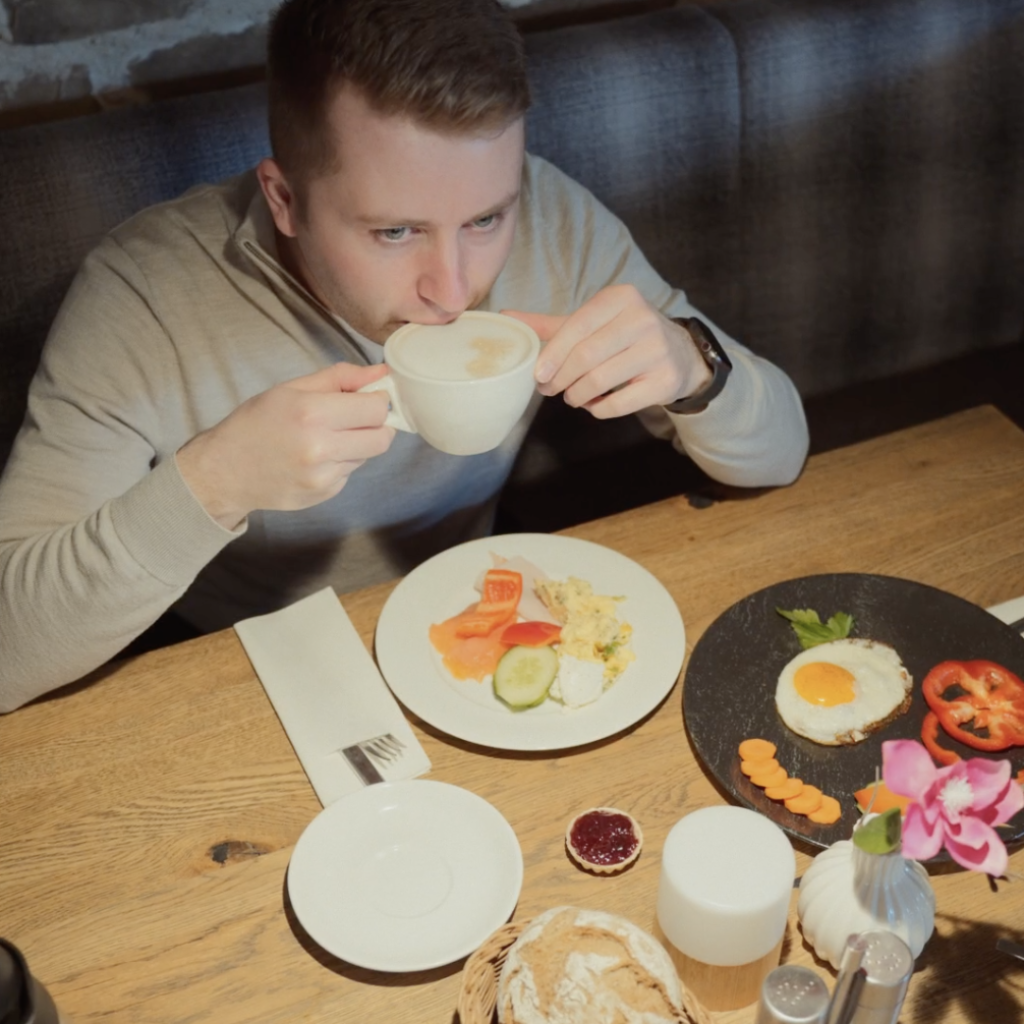 Breakfast at Peter’s - Der perfekte Start in den Tag 9 Ein Mann sitzt an einem Holztisch und nippt aus einer weißen Tasse. Vor ihm stehen Teller mit Frühstücksspeisen, darunter Eier, Gemüse, Brot und ein kleines Glas mit Marmelade. Der Tisch ist gut beleuchtet und ordentlich gedeckt.