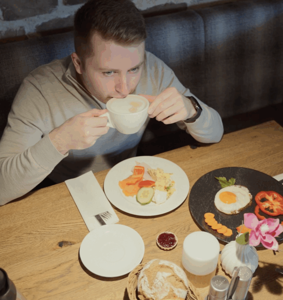 Ein Mann sitzt an einem Holztisch und nippt aus einer weißen Tasse. Vor ihm stehen Teller mit Frühstücksspeisen, darunter Eier, Gemüse, Brot und ein kleines Glas mit Marmelade. Der Tisch ist gut beleuchtet und ordentlich gedeckt.