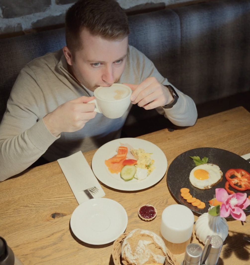 Ein Mann sitzt an einem Holztisch und nippt aus einer weißen Tasse. Vor ihm stehen Teller mit Frühstücksspeisen, darunter Eier, Gemüse, Brot und ein kleines Glas mit Marmelade. Der Tisch ist gut beleuchtet und ordentlich gedeckt.