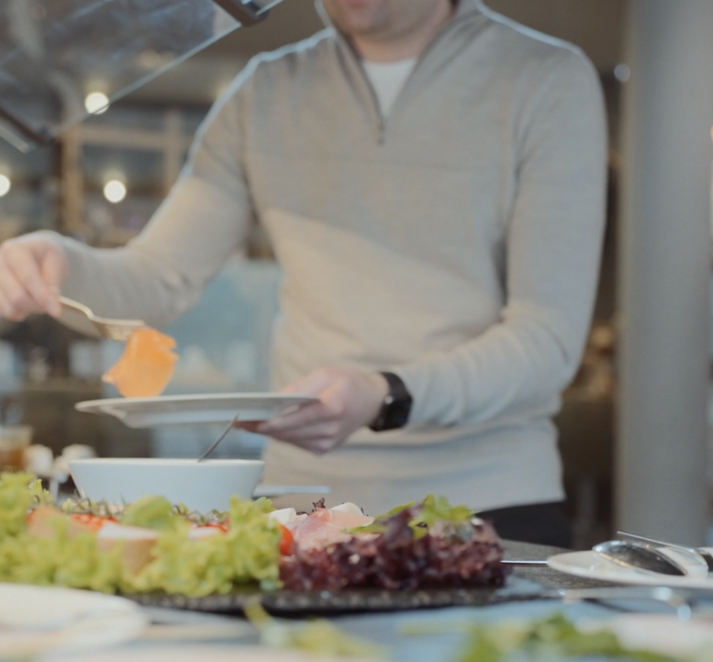 Eine Person in einem hellen Pullover serviert Lachsscheiben auf einem Teller an einem Büffettisch mit verschiedenen Grün- und Salatarten im Vordergrund.