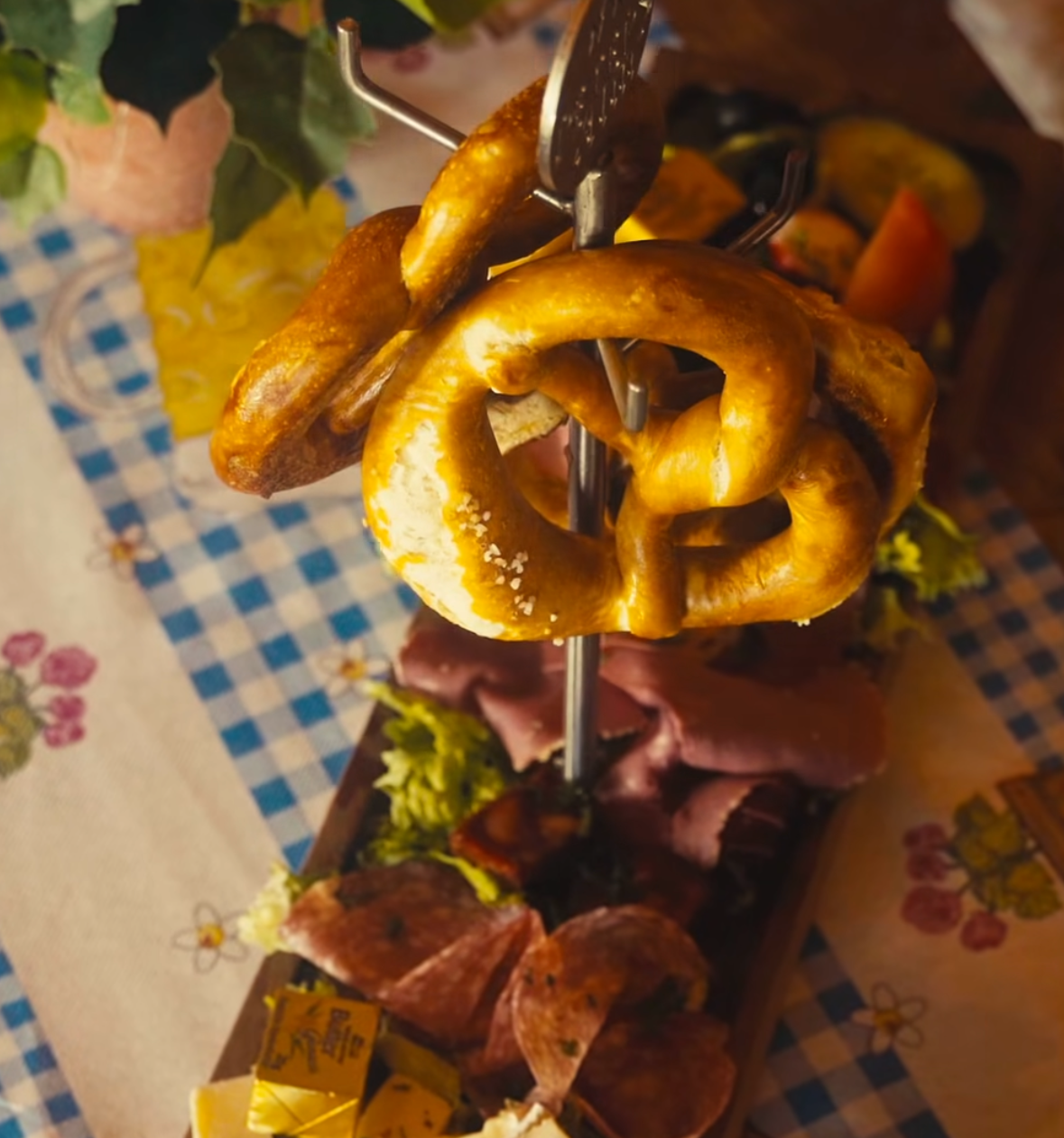 Auf einem Holzbrett auf einer blau-karierten Tischdecke liegen Aufschnitt, Käse, Zwiebelscheiben und Essiggurken, dazu zwei Brezeln auf einem Metallständer, und im Hintergrund ist eine Pflanze zu sehen.