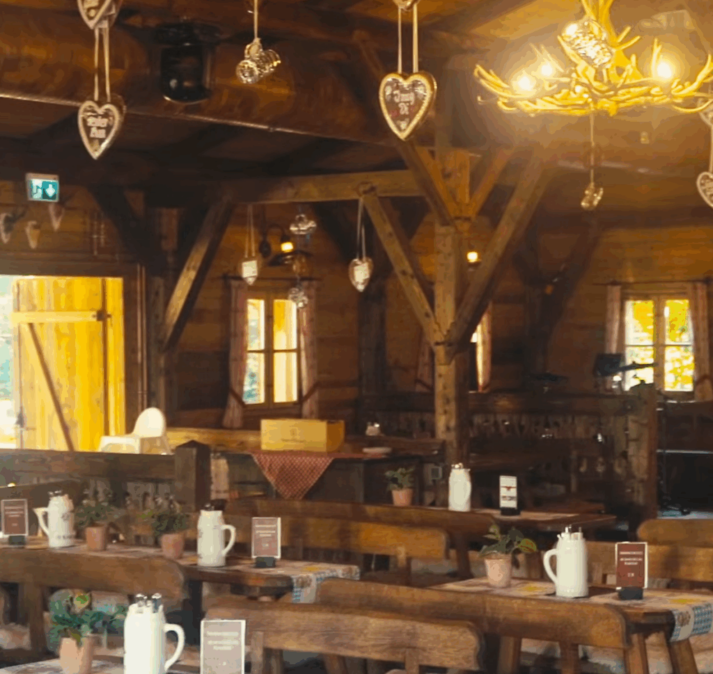Ein gemütliches Holzrestaurant mit rustikalen Tischen und Bänken, weißen Krügen als Tafelaufsatz, von der Decke hängenden herzförmigen Dekorationen und warmer Raumbeleuchtung.
