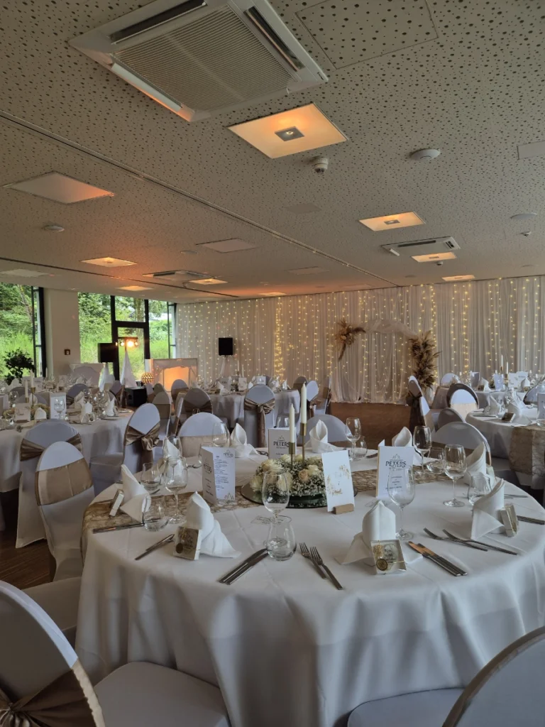 Ein dekorierter Hochzeitssaal mit runden Tischen, die mit weißer Bettwäsche, ordentlich gefalteten Servietten, Gläsern, Tafelaufsätzen und Tischkarten für die Gäste bedeckt sind. Die weißen Stühle haben beigefarbene Schärpen, und an der Rückwand hängen Lichterketten.
