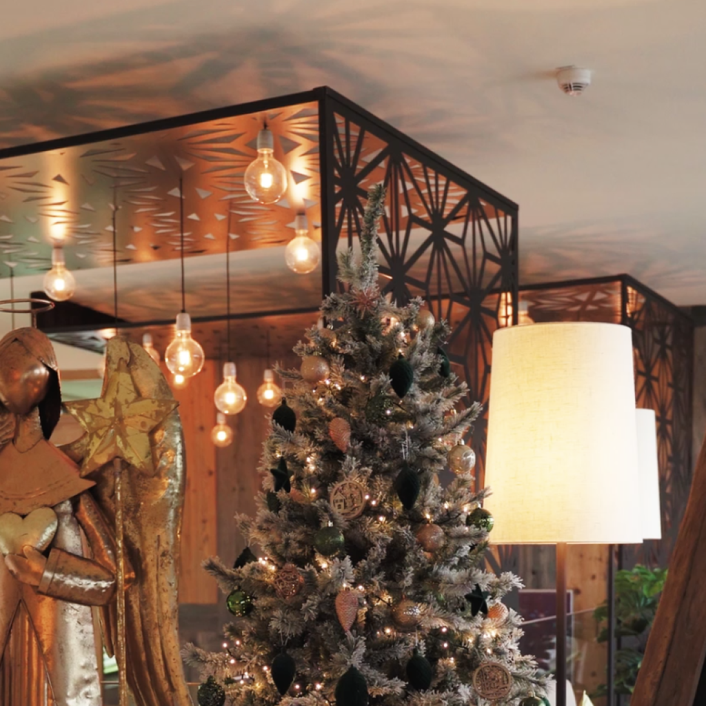 Weihnachten und Silvester – Entdecken Sie die Magie der Festtage neu 6 Ein mit Ornamenten und Lichtern geschmückter Weihnachtsbaum steht neben einer goldenen Engelsfigur in einem warm beleuchteten Raum mit modernem Dekor und hängenden Glühbirnen. Auf der rechten Seite ist teilweise ein Kreideschild zu sehen.