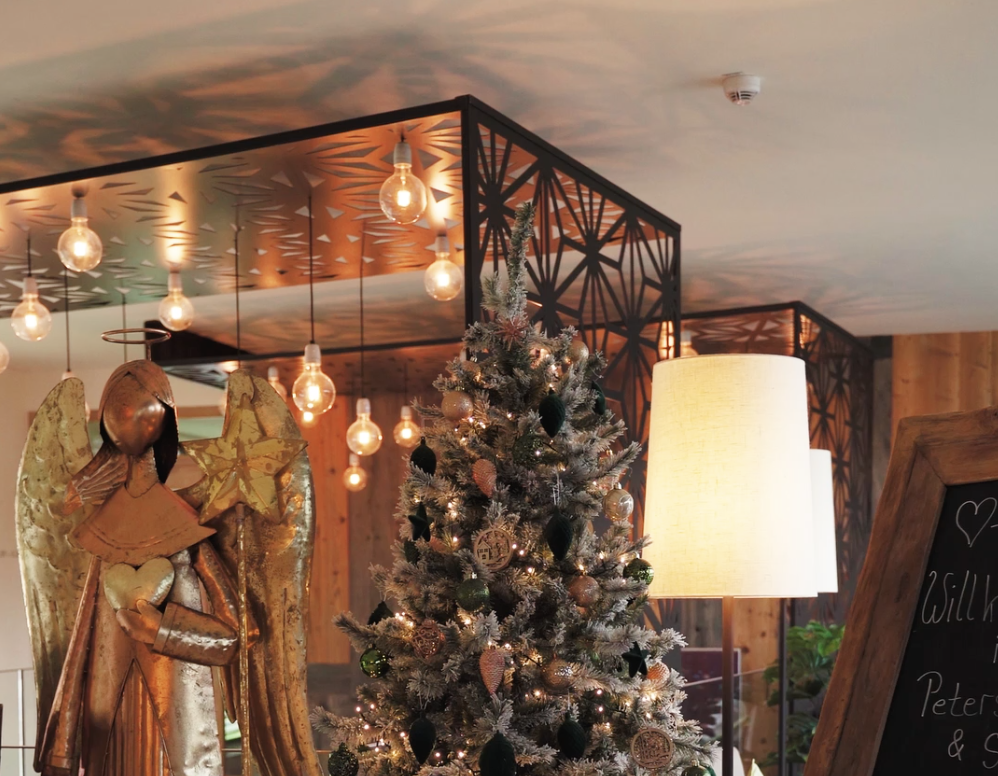 Ein mit Ornamenten und Lichtern geschmückter Weihnachtsbaum steht neben einer goldenen Engelsfigur in einem warm beleuchteten Raum mit modernem Dekor und hängenden Glühbirnen. Auf der rechten Seite ist teilweise ein Kreideschild zu sehen.