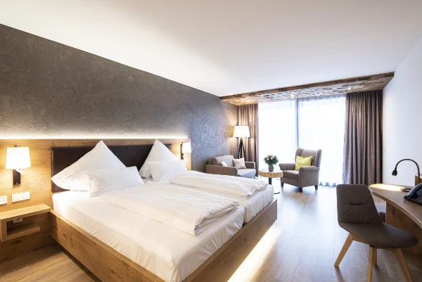 Ein modernes Hotelzimmer mit einem großen Bett, weißer Bettwäsche, Holzmöbeln, einer Sitzecke mit zwei Sesseln, einem kleinen Tisch, Stehlampen, einem Schreibtisch mit Stuhl und großen Fenstern mit Vorhängen, die natürliches Licht hereinlassen.