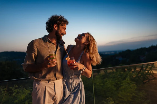 Ein lächelndes Paar steht bei Sonnenuntergang dicht beieinander auf einem Balkon, hält bunte Getränke in der Hand und schaut sich an, während im Hintergrund Grün und ein klarer Himmel zu sehen sind.