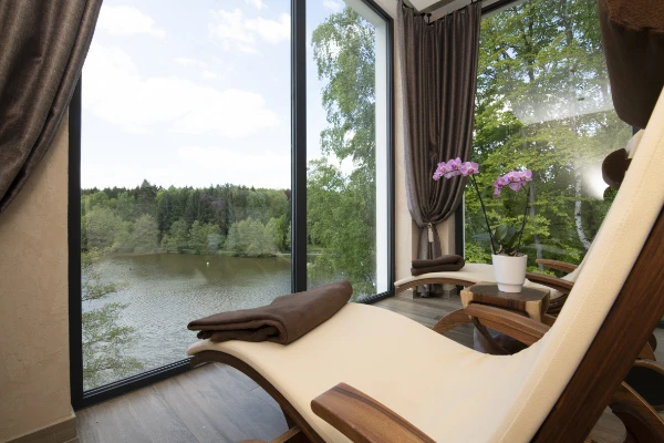 Ein Loungesessel mit einem braunen Handtuch neben einem großen Fenster mit Blick auf einen ruhigen See und üppig grüne Bäume, mit einer Orchideenpflanze auf einem Tisch in der Nähe, schafft eine friedliche, entspannende Atmosphäre.