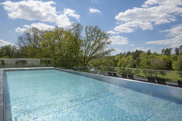Infinity-Pool mit klarem blauen Wasser, umgeben von Liegestühlen und Glasgeländern, mit Blick auf eine üppige grüne Landschaft und Bäume unter einem teilweise bewölkten Himmel.