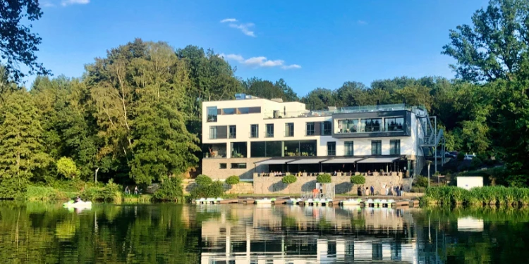Ein modernes weißes Gebäude mit großen Fenstern steht an einem ruhigen See, umgeben von üppigen grünen Bäumen. Sein Spiegelbild ist im Wasser zu sehen, und auf einer Terrasse in Ufernähe sind Menschen versammelt.