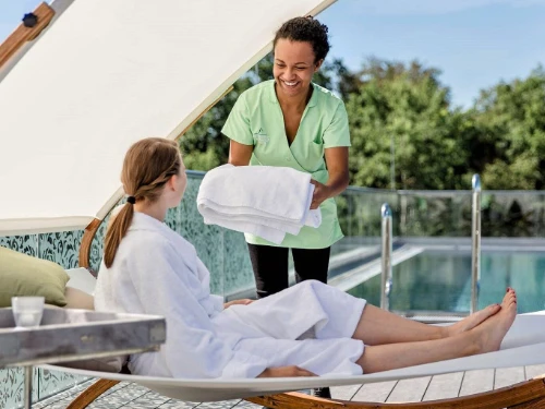 Unser Servicepersonal reicht einer Frau, die sich auf einem Liegestuhl an einem Außenpool entspannt, frische Handtücher. Die Frau trägt einen weißen Bademantel, und beide lächeln. Im Hintergrund sind Bäume zu sehen.