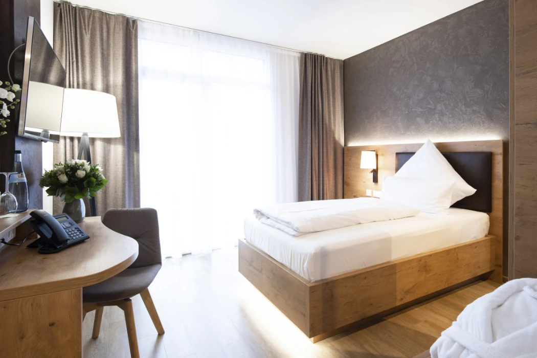 Ein modernes Hotelzimmer mit einem Einzelbett aus Holz, weißem Bettzeug, einem Schreibtisch mit Stuhl, einem Telefon, einer Blumenvase, einem an der Wand montierten Fernseher und großen Fenstern mit durchsichtigen Vorhängen, die natürliches Licht hereinlassen.