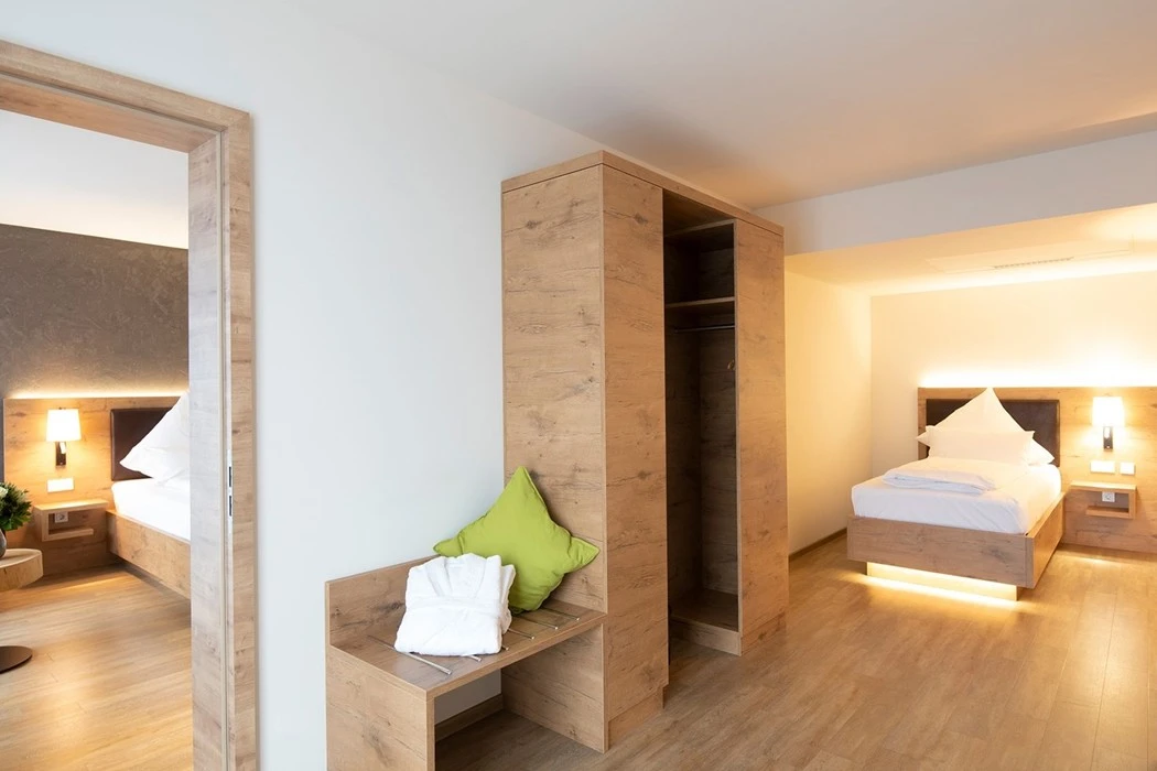 Modernes Hotelzimmer mit zwei Einzelbetten, Holzmöbeln, einem offenen Kleiderschrank, einer Bank mit grünem Kissen und warmer Beleuchtung, die eine gemütliche Atmosphäre schafft. Eine Tür führt in einen anderen Bereich mit einem weiteren Bett.
