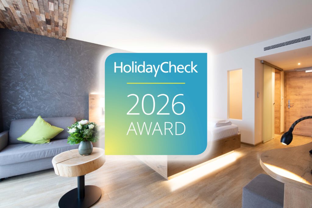HolidayCheck Award 2026: PETERS Hotel & Spa erneut ausgezeichnet 1 Helles, modernes Hotelzimmer mit grauem Sofa, grünem Kissen, Holzmöbeln, Blumen auf einem Tisch und dem HolidayCheck 2026 Award-Logo, das in der Mitte eingeblendet ist.